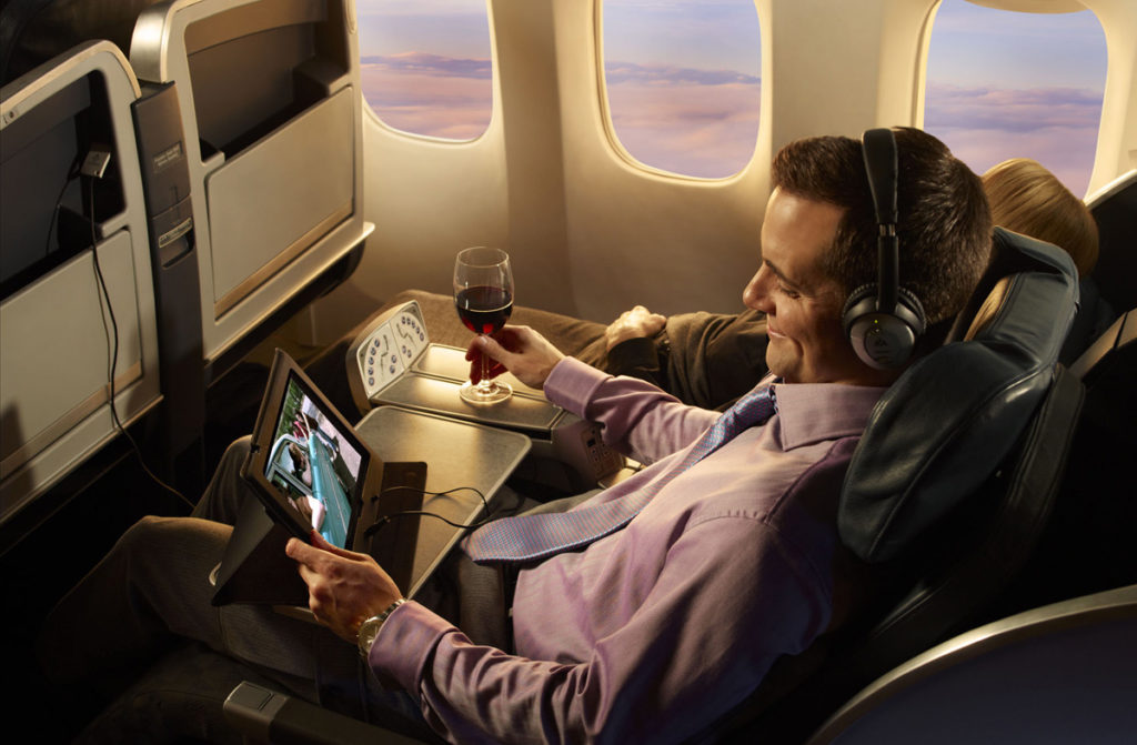 A man watches a movie on his tablet on an airplane while drinking a glass of red wine -American Airlines inflight entertainment is now free! ASAP Tickets Travel Blog