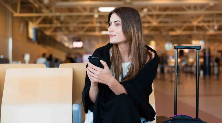 A smiling woman waits at the airport holding her mobile cell phone - Book Last-Minute Flights